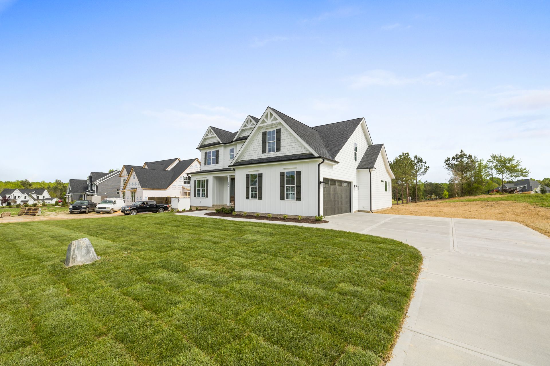 New-construction street with a finished white farmhouse in front and framed houses and work trucks beyond